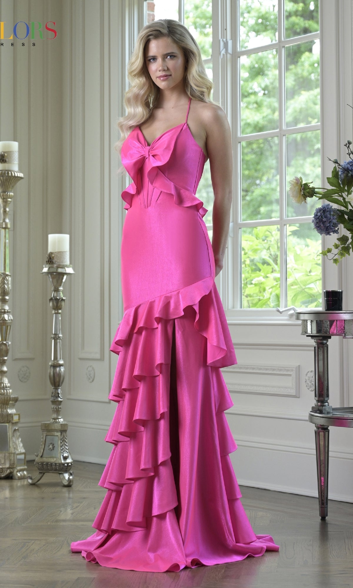 A woman stands indoors by tall windows and elegant decor, wearing the Sheer-Back Ruffle Prom Gown: Colors Dress 3899—a bright pink satin evening gown with ruffles and bow detail, perfect for special occasions.
